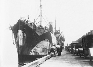 A large ship is docked at a harbor, showing a distinctive black and white camouflage pattern on its hull. Several men stand on the wooden dock near the ship, and a line of vintage vehicles is parked alongside the dock. The background is bright and somewhat overexposed, giving a clear view of the scene.