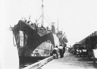 A large ship is docked at a harbor, showing a distinctive black and white camouflage pattern on its hull. Several men stand on the wooden dock near the ship, and a line of vintage vehicles is parked alongside the dock. The background is bright and somewhat overexposed, giving a clear view of the scene.