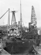 A large ship is docked at a busy industrial port. The scene includes various cranes and cables, with workers attending to the ship. The ship appears to be undergoing maintenance or loading. Several masts and smokestacks are visible, and the area is filled with mechanical infrastructure.