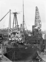 A large ship is docked at a busy industrial port. The scene includes various cranes and cables, with workers attending to the ship. The ship appears to be undergoing maintenance or loading. Several masts and smokestacks are visible, and the area is filled with mechanical infrastructure.