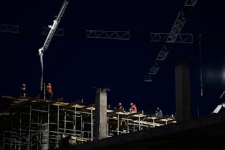 Construction workers are seen working at a building site at night, with scaffolding and cranes visible. The scene is illuminated by construction lights, casting a bright light on the workers wearing safety gear including helmets and reflective vests. The sky is dark, indicating either early morning or late evening.