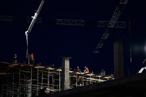 Construction workers are seen working at a building site at night, with scaffolding and cranes visible. The scene is illuminated by construction lights, casting a bright light on the workers wearing safety gear including helmets and reflective vests. The sky is dark, indicating either early morning or late evening.