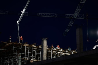 Construction workers are seen working at a building site at night, with scaffolding and cranes visible. The scene is illuminated by construction lights, casting a bright light on the workers wearing safety gear including helmets and reflective vests. The sky is dark, indicating either early morning or late evening.