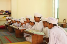 A group of young boys in white traditional attire are seated on the floor, intently reading from open books placed on low wooden tables in a classroom setting with muted colors and some books shelved on the side.