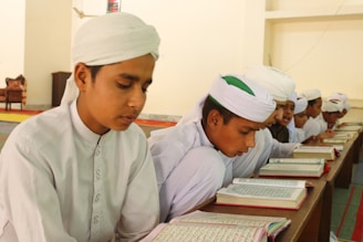 Young boys wearing traditional white attire and turbans are sitting together in a classroom setting. They are focused on reading and studying from open books placed on a wooden desk. The environment appears to be a learning or religious study setting with simple, neutral walls and a carpeted floor.