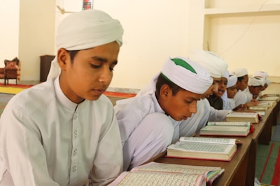 Young boys wearing traditional white attire and turbans are sitting together in a classroom setting. They are focused on reading and studying from open books placed on a wooden desk. The environment appears to be a learning or religious study setting with simple, neutral walls and a carpeted floor.