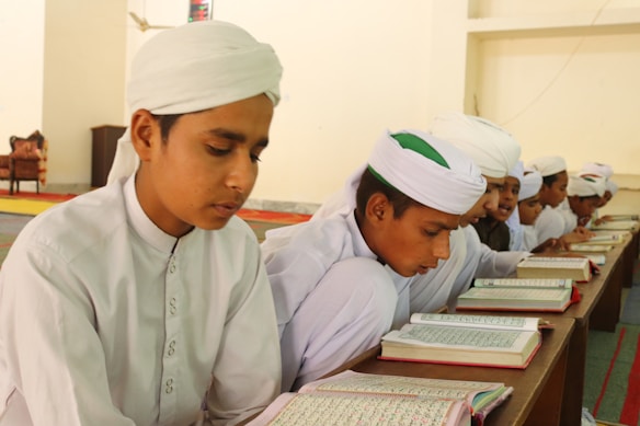 Young boys wearing traditional white attire and turbans are sitting together in a classroom setting. They are focused on reading and studying from open books placed on a wooden desk. The environment appears to be a learning or religious study setting with simple, neutral walls and a carpeted floor.