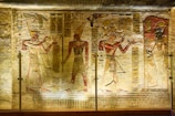 Ancient Egyptian wall carvings featuring three figures in traditional attire, adorned with headdresses, and holding various objects. Hieroglyphics and symbolic images are intricately carved around the figures, set against a slightly worn stone background. The lighting highlights the detailed artwork and enhances the colors of the carvings.