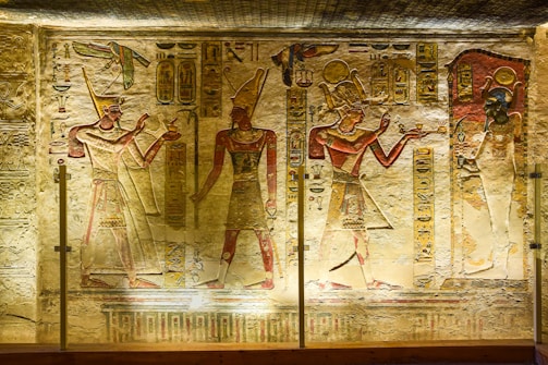 Ancient Egyptian wall carvings featuring three figures in traditional attire, adorned with headdresses, and holding various objects. Hieroglyphics and symbolic images are intricately carved around the figures, set against a slightly worn stone background. The lighting highlights the detailed artwork and enhances the colors of the carvings.