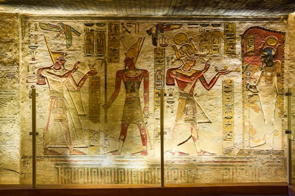 Ancient Egyptian wall carvings featuring three figures in traditional attire, adorned with headdresses, and holding various objects. Hieroglyphics and symbolic images are intricately carved around the figures, set against a slightly worn stone background. The lighting highlights the detailed artwork and enhances the colors of the carvings.