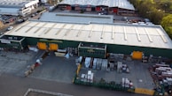 An aerial view of a large industrial warehouse with a green exterior and orange loading bays. The surrounding area includes parking lots filled with parked vehicles and additional storage and loading spaces. The warehouse is situated in a business park environment with other similar buildings nearby.