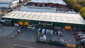 An aerial view of a large industrial warehouse with a green exterior and orange loading bays. The surrounding area includes parking lots filled with parked vehicles and additional storage and loading spaces. The warehouse is situated in a business park environment with other similar buildings nearby.