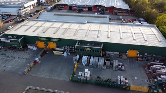 An aerial view of a large industrial warehouse with a green exterior and orange loading bays. The surrounding area includes parking lots filled with parked vehicles and additional storage and loading spaces. The warehouse is situated in a business park environment with other similar buildings nearby.