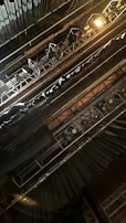 The image depicts an overhead view of stage lighting rigging, featuring multiple spotlights and hanging lights. The scene is set in a theater or concert space, with visible metal structures supporting the lighting equipment. The atmosphere suggests technical preparation for a performance.