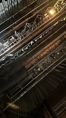 The image depicts an overhead view of stage lighting rigging, featuring multiple spotlights and hanging lights. The scene is set in a theater or concert space, with visible metal structures supporting the lighting equipment. The atmosphere suggests technical preparation for a performance.