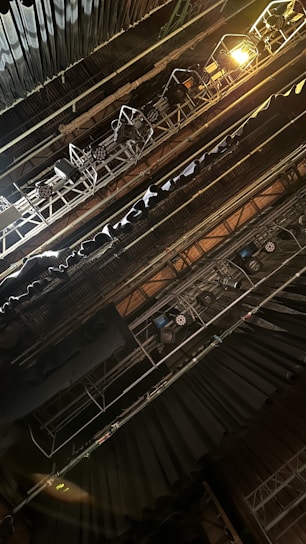 The image depicts an overhead view of stage lighting rigging, featuring multiple spotlights and hanging lights. The scene is set in a theater or concert space, with visible metal structures supporting the lighting equipment. The atmosphere suggests technical preparation for a performance.
