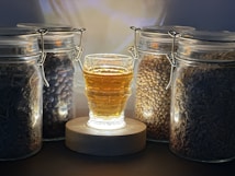 A glass container filled with a golden liquid is positioned in the center on a wooden base, surrounded by five sealed glass jars containing different types of grains, seeds, and other dried materials.