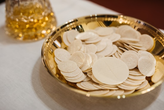 A gold-colored bowl containing a stack of round white communion wafers, placed on a cream-colored tablecloth. In the background, there is a glass container filled with a golden liquid.