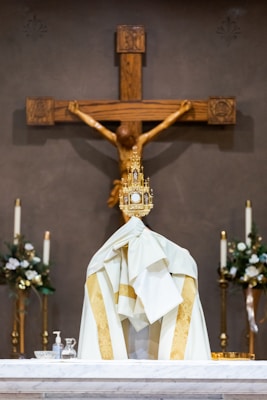 A religious setting featuring a large wooden crucifix with a figure of Jesus at the center. In the foreground, there is a draped white and gold cloth covering a table, with a golden monstrance placed on top. Two lit candles and floral arrangements can be seen on either side, adding to the solemn atmosphere.