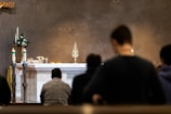 A religious ceremony is taking place with people kneeling in prayer before an altar. The altar is adorned with ornate candlesticks, a golden monstrance, chalices, and floral decorations. Soft lighting casts a reverential atmosphere over the setting.