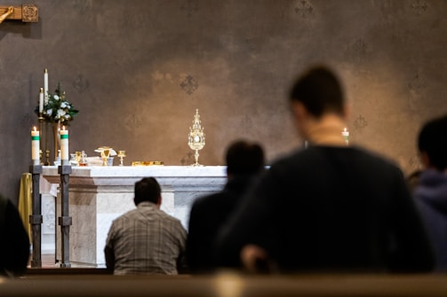 A religious ceremony is taking place with people kneeling in prayer before an altar. The altar is adorned with ornate candlesticks, a golden monstrance, chalices, and floral decorations. Soft lighting casts a reverential atmosphere over the setting.