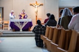People are gathered inside a church, participating in a religious service. Two clergy members stand at the altar, which is decorated with a purple cloth and a display of wheat. Congregants are seated or kneeling in wooden pews, facing the altar. A crucifix is mounted on the wall behind the altar.