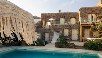A rustic stone villa with multiple windows is complemented by a clear swimming pool in the foreground. The villa features a staircase leading up to an arched balcony, and is surrounded by lush greenery. A straw fringe parasol is partially visible, adding to the tropical ambiance.