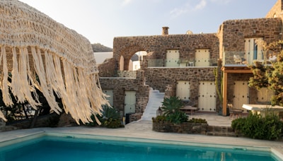 A rustic stone villa with multiple windows is complemented by a clear swimming pool in the foreground. The villa features a staircase leading up to an arched balcony, and is surrounded by lush greenery. A straw fringe parasol is partially visible, adding to the tropical ambiance.