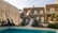 A rustic stone villa with multiple windows is complemented by a clear swimming pool in the foreground. The villa features a staircase leading up to an arched balcony, and is surrounded by lush greenery. A straw fringe parasol is partially visible, adding to the tropical ambiance.
