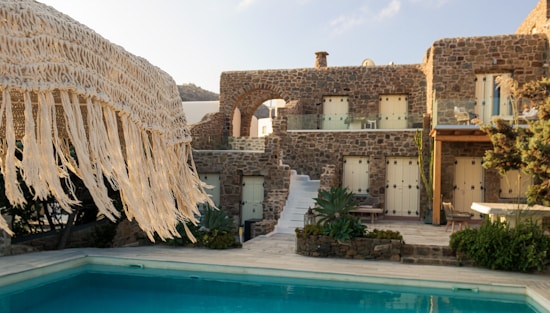A rustic stone villa with multiple windows is complemented by a clear swimming pool in the foreground. The villa features a staircase leading up to an arched balcony, and is surrounded by lush greenery. A straw fringe parasol is partially visible, adding to the tropical ambiance.