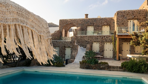 A rustic stone villa with multiple windows is complemented by a clear swimming pool in the foreground. The villa features a staircase leading up to an arched balcony, and is surrounded by lush greenery. A straw fringe parasol is partially visible, adding to the tropical ambiance.