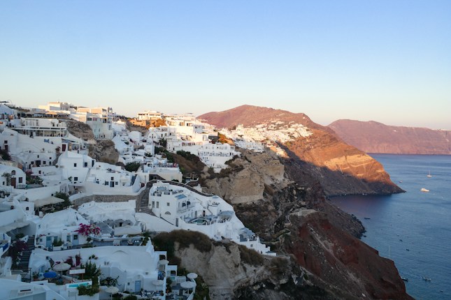 A scenic view of whitewashed buildings built on a steep hillside overlooking a calm blue sea. The architecture is characteristic of a coastal Mediterranean town, with the sun setting and casting a warm glow over the cliffs and buildings.