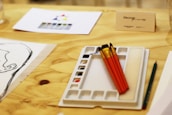 A wooden table holds art supplies featuring three paintbrushes with red handles resting on a white palette with multiple small compartments. Nearby, there is a pencil, a sketch of a head, and a paper with a color wheel. A card with text 'Discover the artist' is placed on the table.