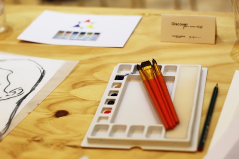 A wooden table holds art supplies featuring three paintbrushes with red handles resting on a white palette with multiple small compartments. Nearby, there is a pencil, a sketch of a head, and a paper with a color wheel. A card with text 'Discover the artist' is placed on the table.
