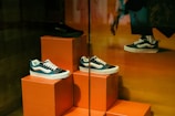 A display of three sneakers is arranged on orange pedestals inside a store. The shoes are a mix of black, white, and teal, with white laces, and they are positioned at different heights. Part of a person's leg and hand, possibly belonging to a mannequin, is visible in the upper right corner, suggesting a retail environment.