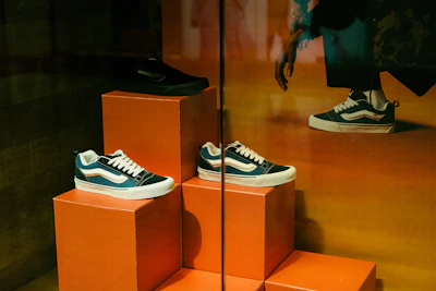 A display of three sneakers is arranged on orange pedestals inside a store. The shoes are a mix of black, white, and teal, with white laces, and they are positioned at different heights. Part of a person's leg and hand, possibly belonging to a mannequin, is visible in the upper right corner, suggesting a retail environment.