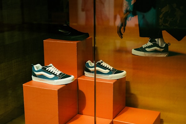 A display of three sneakers is arranged on orange pedestals inside a store. The shoes are a mix of black, white, and teal, with white laces, and they are positioned at different heights. Part of a person's leg and hand, possibly belonging to a mannequin, is visible in the upper right corner, suggesting a retail environment.