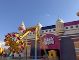 A colorful amusement park ride named Frenzy Station is prominently displayed. The ride features a bright yellow and magenta swinging arm with multiple seats attached, set against the backdrop of a whimsical building with towers and domes reminiscent of a castle. The entrance to the ride has a large pink sign with the name in bold white letters.