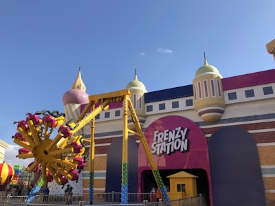 A colorful amusement park ride named Frenzy Station is prominently displayed. The ride features a bright yellow and magenta swinging arm with multiple seats attached, set against the backdrop of a whimsical building with towers and domes reminiscent of a castle. The entrance to the ride has a large pink sign with the name in bold white letters.