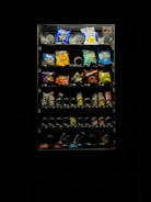 A dimly lit vending machine filled with various snack items, including chips and candy. The image features bright packs in a range of colors such as blue, red, and yellow. The vending machine is surrounded by darkness, emphasizing its illuminated display.