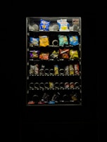 A dimly lit vending machine filled with various snack items, including chips and candy. The image features bright packs in a range of colors such as blue, red, and yellow. The vending machine is surrounded by darkness, emphasizing its illuminated display.