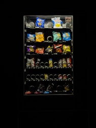 A dimly lit vending machine filled with various snack items, including chips and candy. The image features bright packs in a range of colors such as blue, red, and yellow. The vending machine is surrounded by darkness, emphasizing its illuminated display.