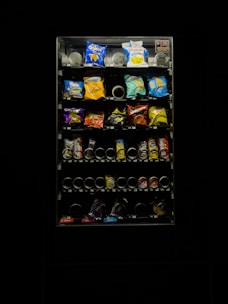 A dimly lit vending machine filled with various snack items, including chips and candy. The image features bright packs in a range of colors such as blue, red, and yellow. The vending machine is surrounded by darkness, emphasizing its illuminated display.
