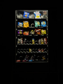 A dimly lit vending machine filled with various snack items, including chips and candy. The image features bright packs in a range of colors such as blue, red, and yellow. The vending machine is surrounded by darkness, emphasizing its illuminated display.
