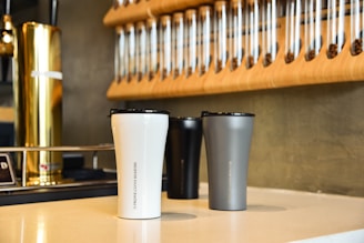 Three insulated travel mugs are placed on a counter in a coffee shop setting. The mugs are white, gray, and black with sleek designs. Behind them, there is a wall-mounted rack holding multiple glass tubes filled with coffee beans. A metallic coffee tap adds a touch of elegance to the scene.