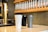 Three insulated travel mugs are placed on a counter in a coffee shop setting. The mugs are white, gray, and black with sleek designs. Behind them, there is a wall-mounted rack holding multiple glass tubes filled with coffee beans. A metallic coffee tap adds a touch of elegance to the scene.
