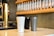 Three insulated travel mugs are placed on a counter in a coffee shop setting. The mugs are white, gray, and black with sleek designs. Behind them, there is a wall-mounted rack holding multiple glass tubes filled with coffee beans. A metallic coffee tap adds a touch of elegance to the scene.