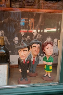 Three caricature figurines with exaggerated features are placed on a shelf in front of a black-and-white photograph of several people. The figures include two men in suits and a woman in a dress, each with distinct and exaggerated facial expressions. Reflections on the glass surface indicate the photo was taken through a window, showing the photographer's silhouette.