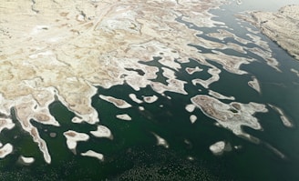 An aerial view of a large water body with irregularly shaped land masses creating a striking pattern. The land appears arid with few vegetation patches, contrasting against the deep green of the water. The image captures the intricate boundary where land meets water, highlighting the unique natural formations.