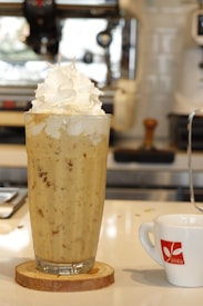 A large glass filled with a creamy, frothy beverage topped with a generous amount of whipped cream stands on a wooden coaster. The background reveals parts of a modern coffee machine, adding a café ambiance. A small, white coffee cup with a red logo is positioned to the right.